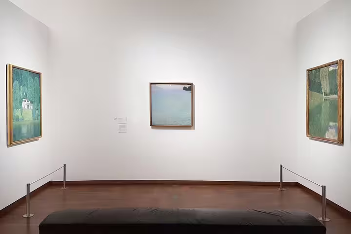 Interior of a gallery room at Belvedere Museum in Vienna showcasing Gustav Klimt paintings on white walls.