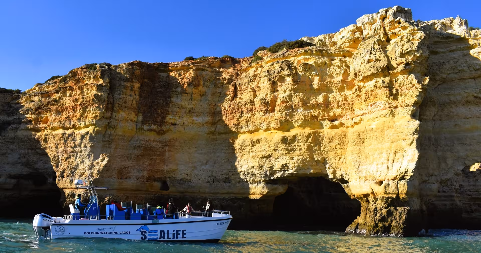 Benagil boat tour cruising past towering golden Algarve sea cliffs and caves under a clear blue sky in southern Portugal