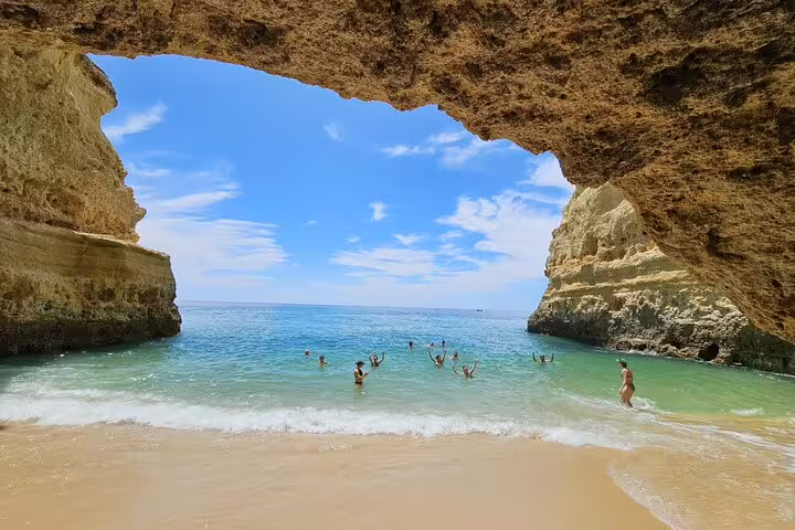 Visitors enjoying the sun and sea at the sandy beach inside Benagil Cave, with clear skies overhead.