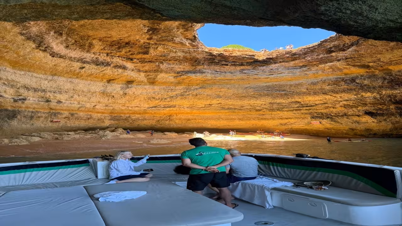 Guests relaxing on a boat inside Benagil Cave, watching kayakers land on the golden Algarve beach beneath the skylight