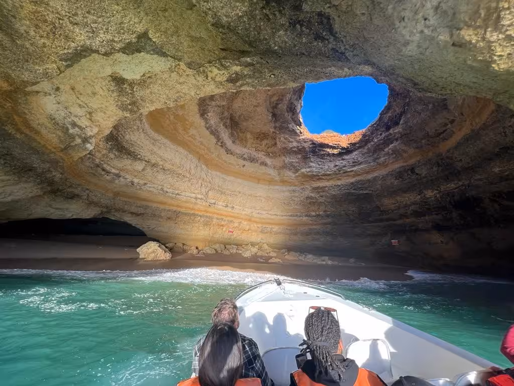 Visitors on a Benagil Cave boat tour glide over turquoise Algarve water toward the sunlit sea cave dome and sandy shore.