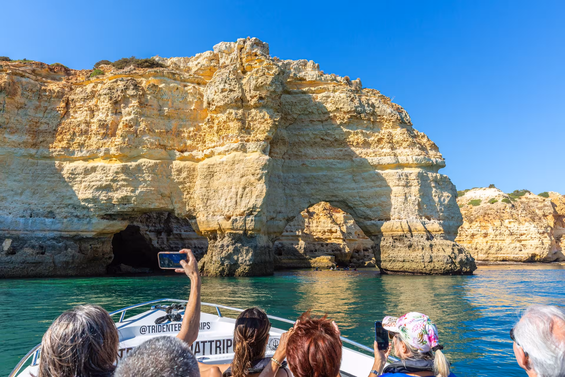 Visitors on a Benagil Cave boat tour photographing dramatic Algarve sea arches and limestone cliffs under clear blue sky