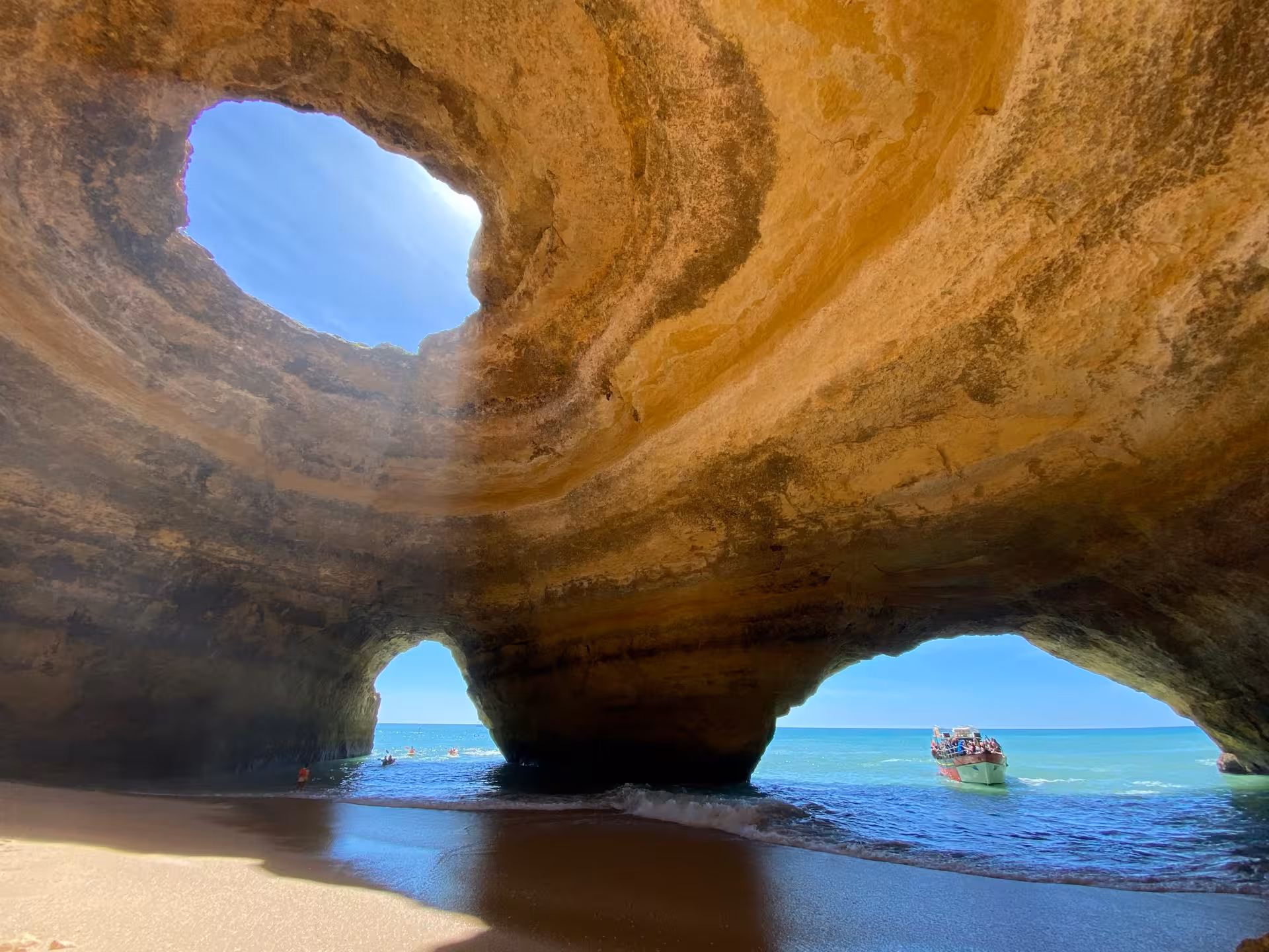 Sunlight streaming through the Benagil Cave skylight onto sandy beach, with tour boat and kayakers at the Algarve coast