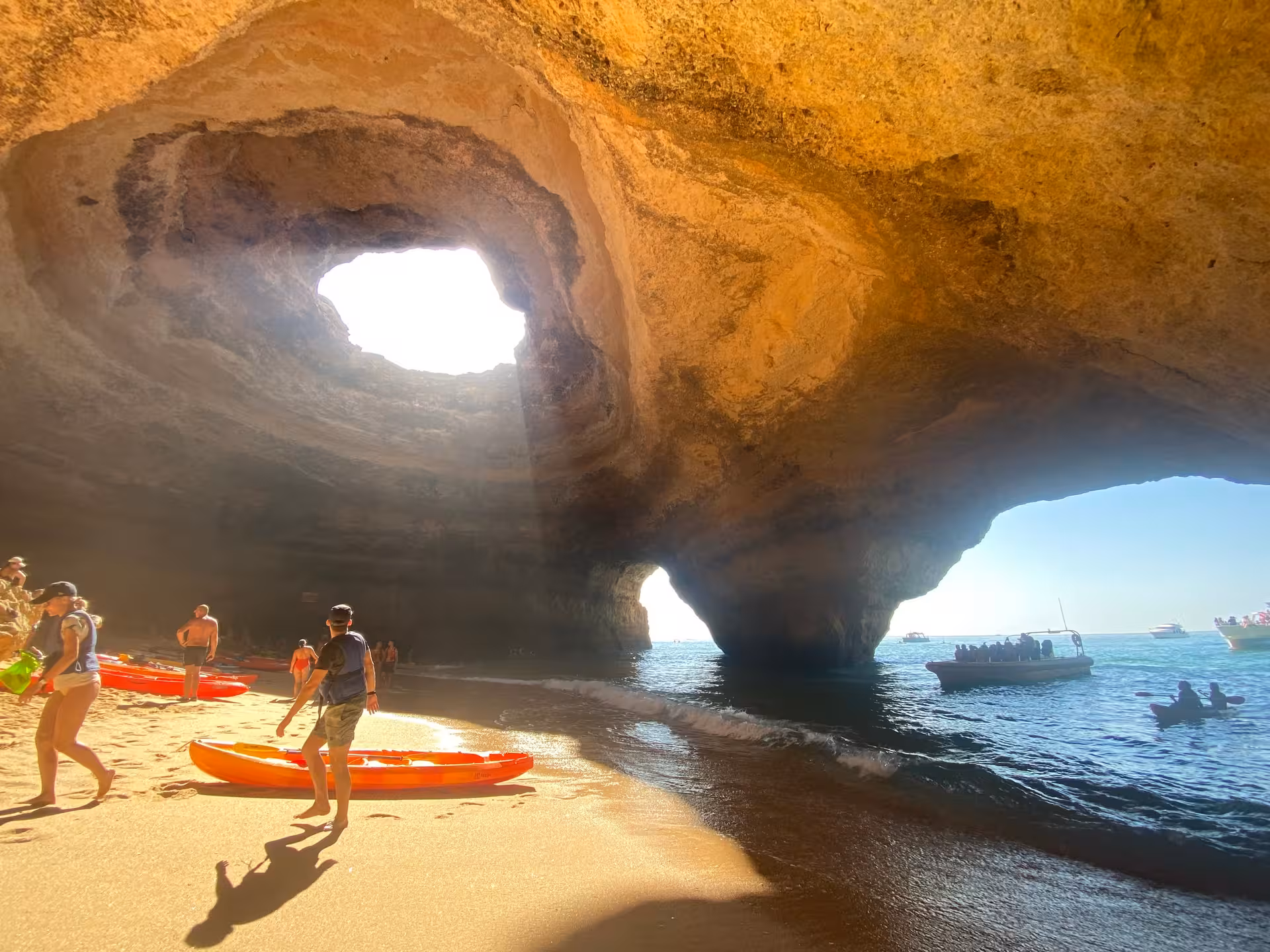 Visitors arriving by kayak and boat at Benagil Cave beach in Algarve, Portugal, with sunlight streaming through rock dome