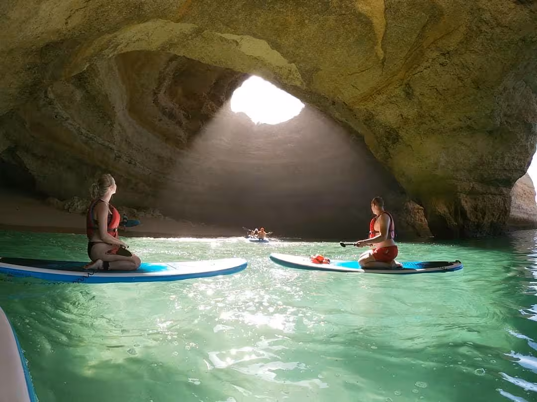 Small group kneels on paddleboards under sunbeam streaming through Benagil Cave ceiling on Algarve stand up paddle tour