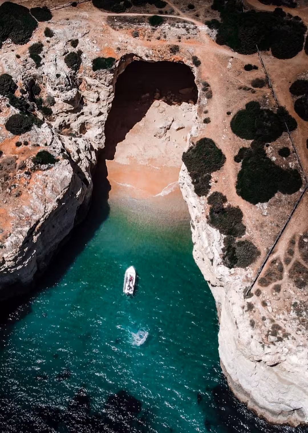 Aerial view of small tour boat approaching secluded Benagil sea cave beach along rugged Algarve coast in Portugal