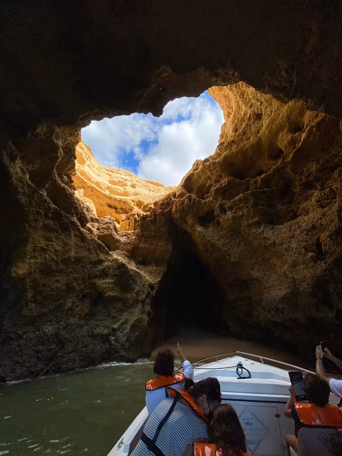 Tour boat gliding into dark Benagil sea cave in Algarve as visitors look up at the dramatic skylight opening and cliffs above