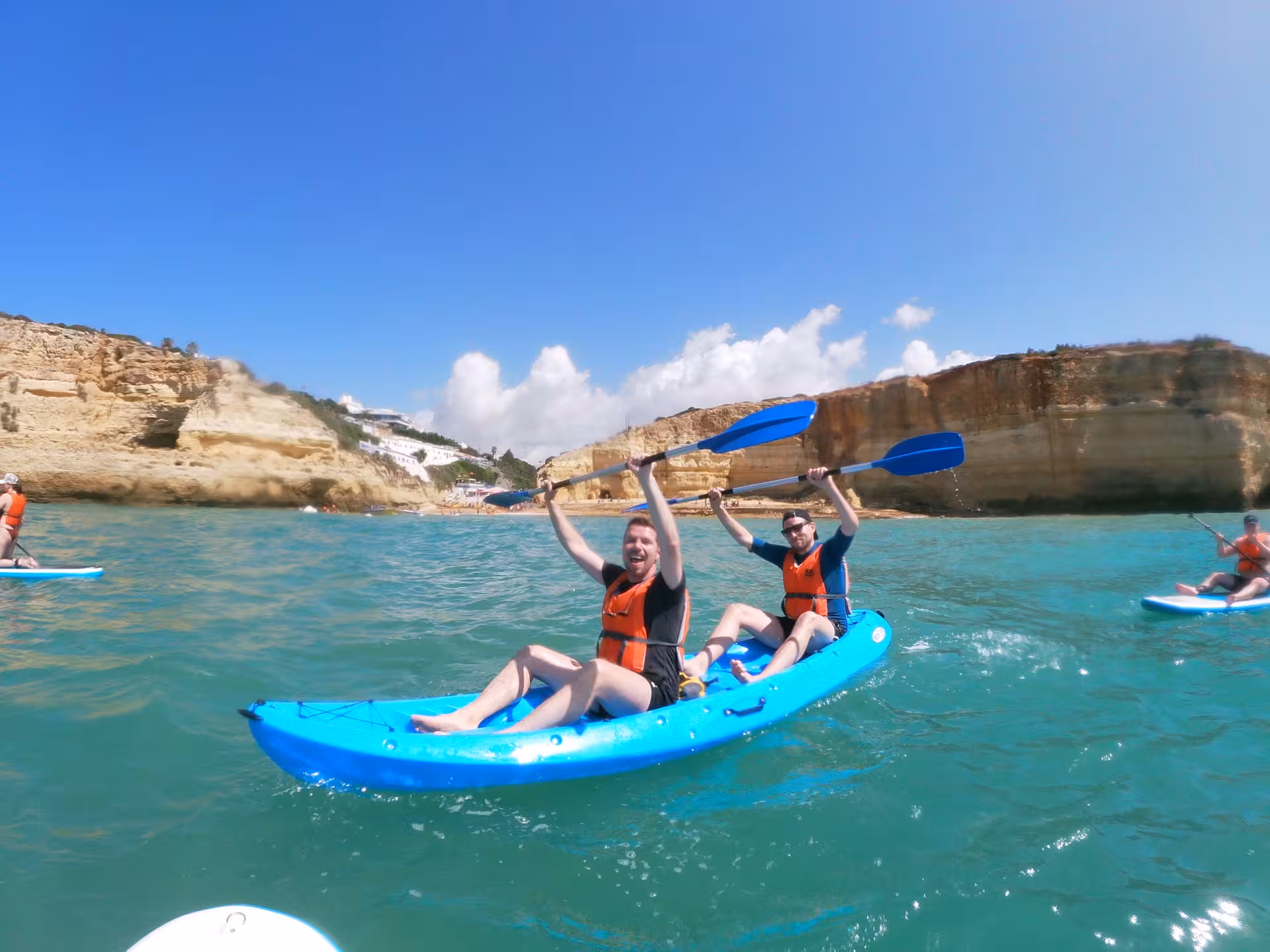 Happy friends on Benagil Caves kayak adventure raising paddles above turquoise Algarve waters with golden cliffs and beach behind