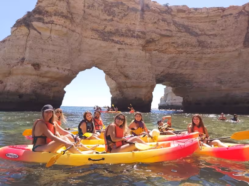 Group sea kayak tour paddling past a dramatic coastal rock arch and caves on a sunny ocean adventure