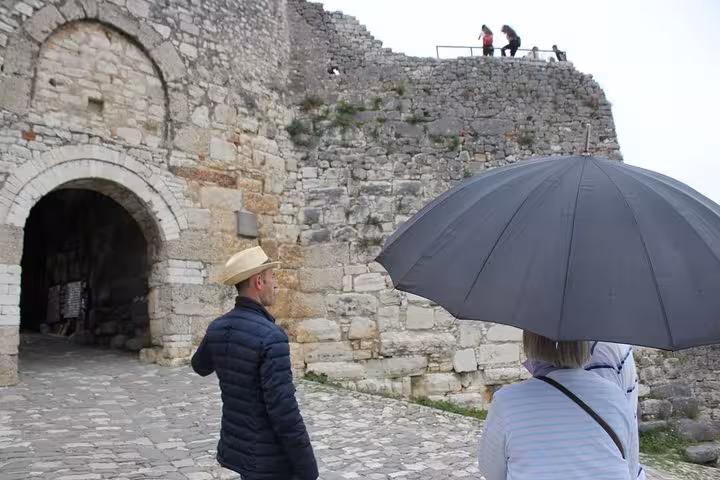 Tour participants admire the ancient stone walls of Berat Castle, sheltered by umbrellas on a cloudy day tour.