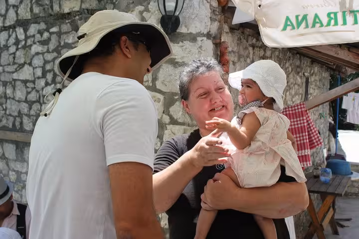 Smiling woman holding a baby, engaging with a man in a sun hat at a Berat cooking class in Albania.
