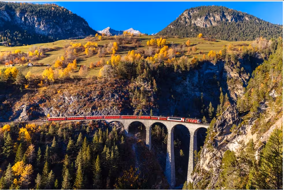 Scenic view of the Bernina Red Train crossing a viaduct amidst vibrant autumn foliage near St Moritz on a clear day.