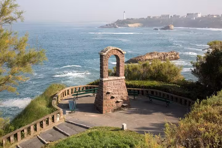 Scenic view of Biarritz coastline with a stone arch, lush greenery, and distant lighthouse under a clear sky.