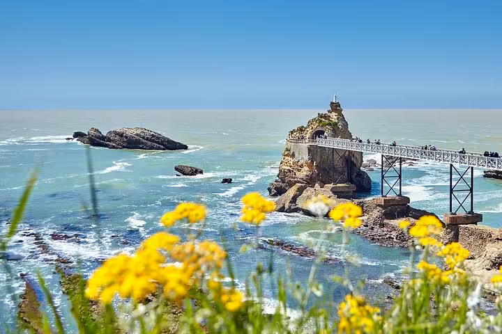 Picturesque view of Biarritz's Rocher de la Vierge with yellow flowers and blue ocean backdrop.