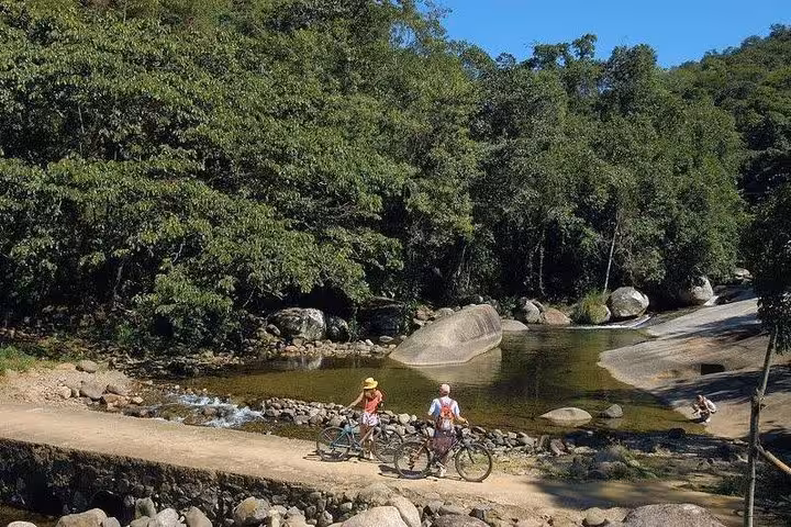 Two cyclists pause by a scenic river in Paraty, surrounded by lush greenery and natural rock formations.