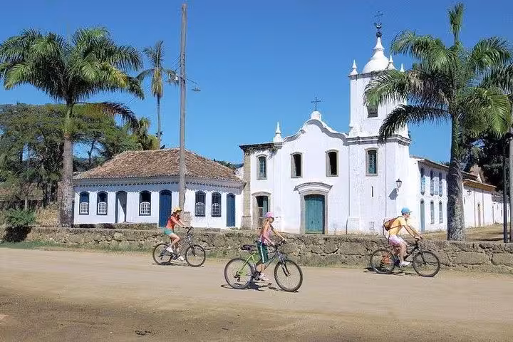 Cyclists explore historic Paraty on bike rentals, passing a charming colonial church under palm trees.