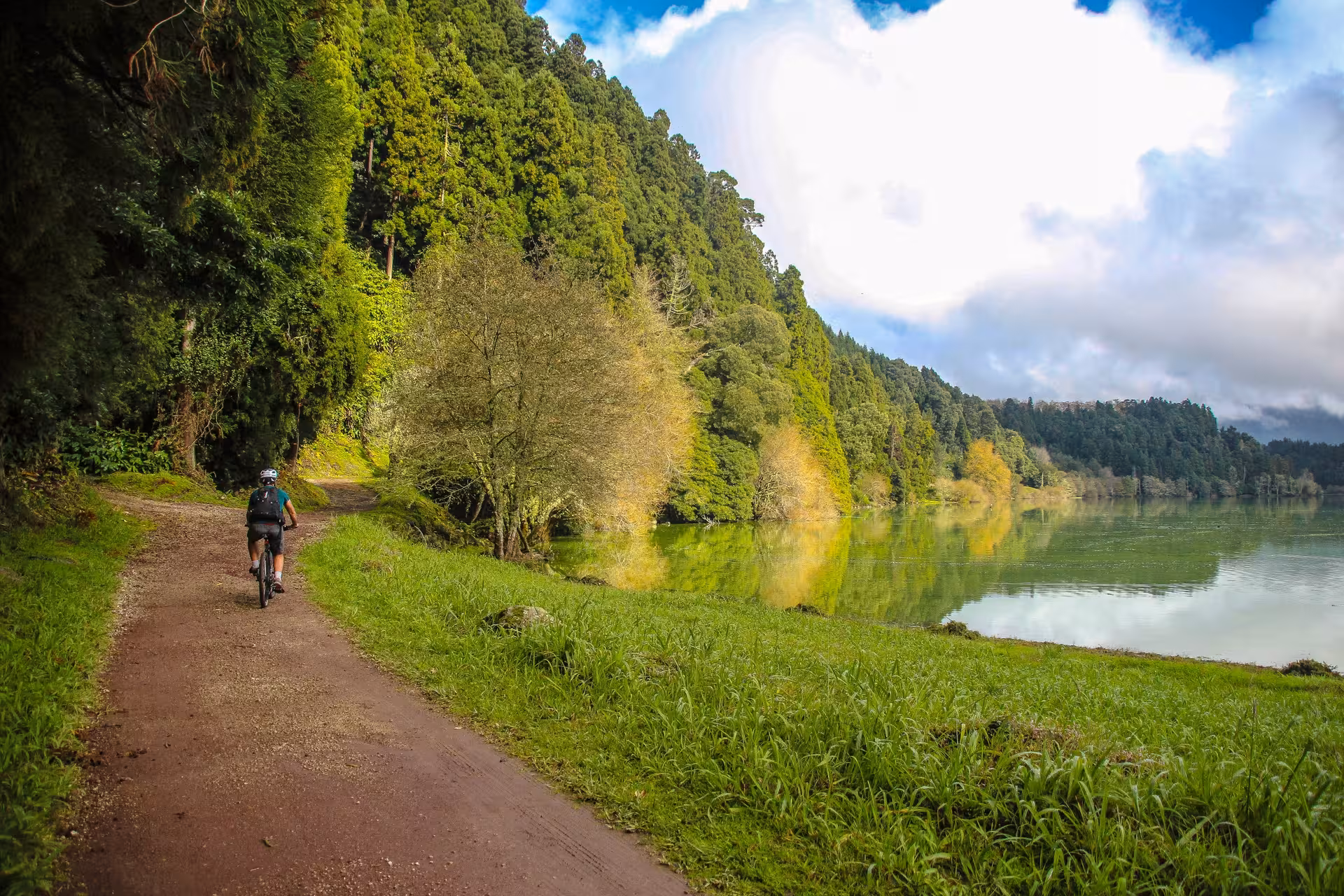Solo cyclist follows a dirt track along the tranquil Sete Cidades lake, exploring lush Azores landscapes by rental bike.