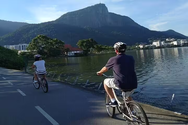 Cyclists enjoy a leisurely ride beside Rio's lagoon with stunning mountain views under a sunny sky.
