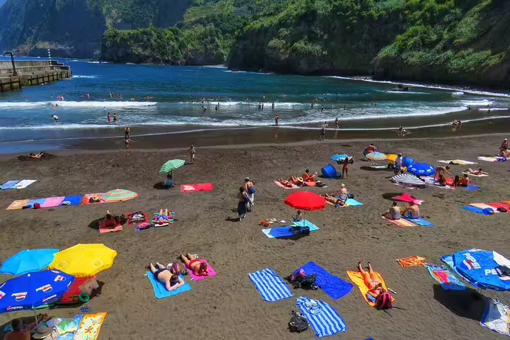 Beachgoers relax on a scenic black sand beach with lush green cliffs, a highlight of the Northwest Terraces and Volcanic Pools 4x4 tour.