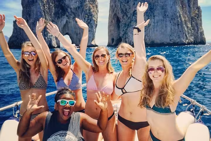 Excited group of friends on a boat tour, enjoying the sunny Amalfi coast with iconic Capri rock formations in view.