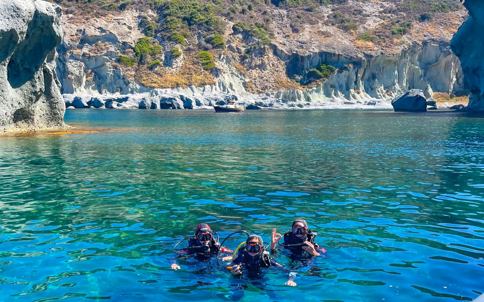 Three divers in full gear explore the crystal-clear waters of Bosa, Sardinia, surrounded by stunning rocky cliffs.