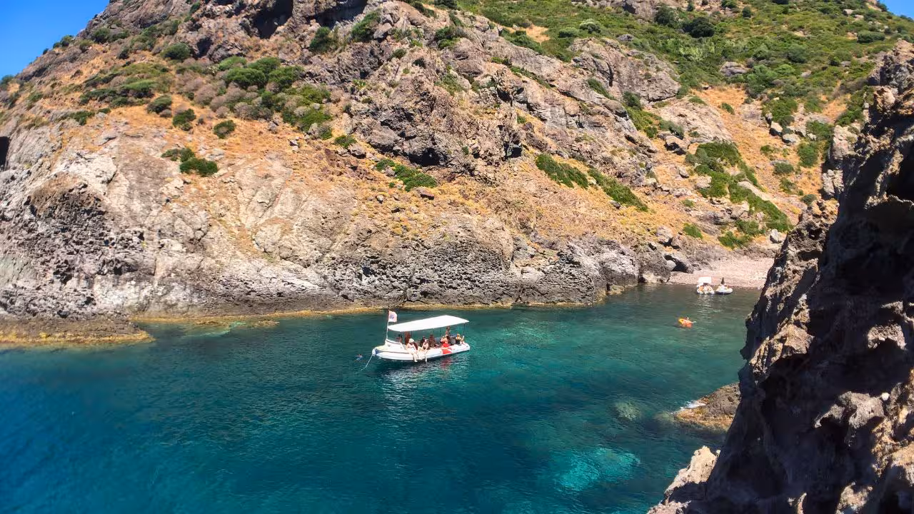 A small boat with tourists navigating the vibrant blue waters of Capo Marrargiu against rugged cliffs in Bosa.