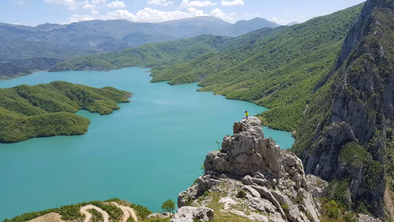 Breathtaking view of Bovilla Lake with lush greenery and a lone adventurer atop a rocky cliff under a bright sky.