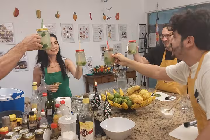 Group of people toasting with caipirinhas in a lively kitchen setting, featuring ingredients for a Brazilian cooking class.