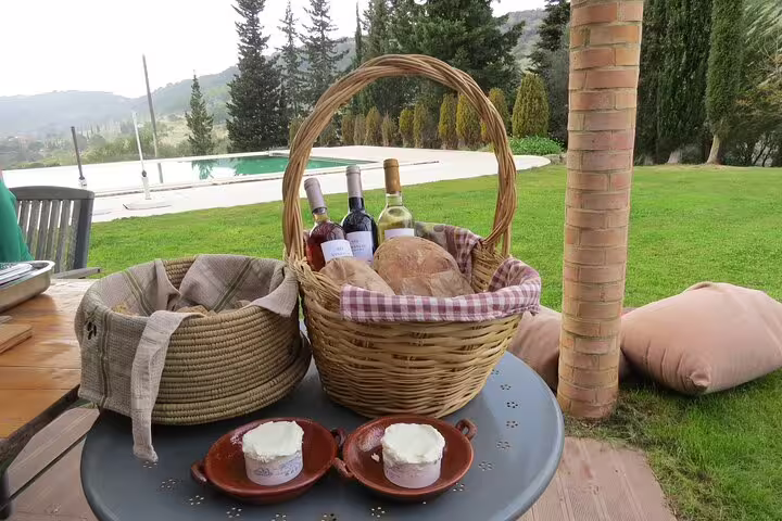 Outdoor table with baskets of bread, wine bottles, and cheese, set against a lush garden and pool backdrop.