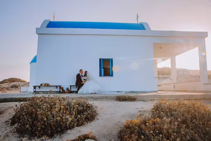 Bride and groom seated by a charming white chapel with blue accents, captured beautifully in Ayia Napa's golden hour.