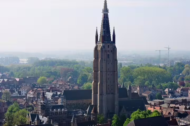 Aerial view of Bruges featuring the iconic Church of Our Lady tower, surrounded by picturesque medieval buildings and lush greenery.