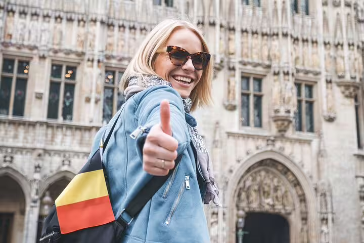 Smiling tourist with Belgian flag backpack gives thumbs up in front of Brussels' historic architecture on a private city tour.