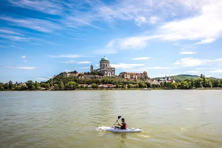 A kayaker paddles on the Danube River with the iconic Esztergom Basilica in the background under a clear blue sky.