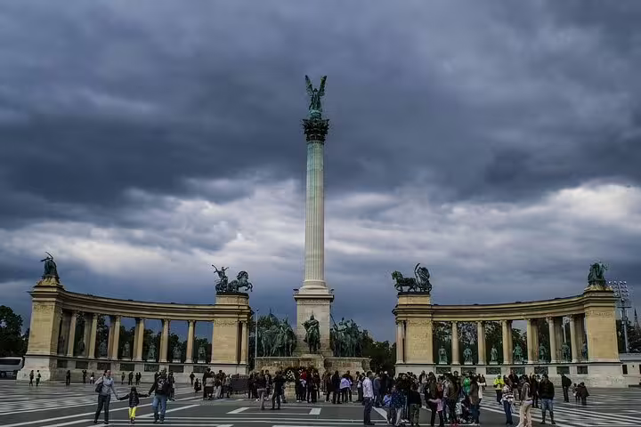 Tourists gather at Budapest's Heroes' Square under dramatic evening skies, highlighting iconic statues and historic architecture.