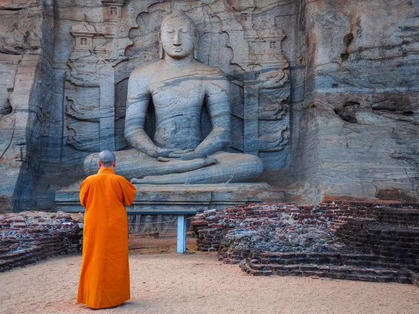 A Buddhist monk in orange robes stands before a large ancient Buddha statue carved into a Sri Lankan rock face.