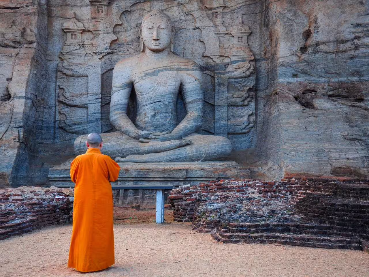 Buddhist monk in orange robes meditating before an ancient rock-carved Buddha statue in Sri Lanka.