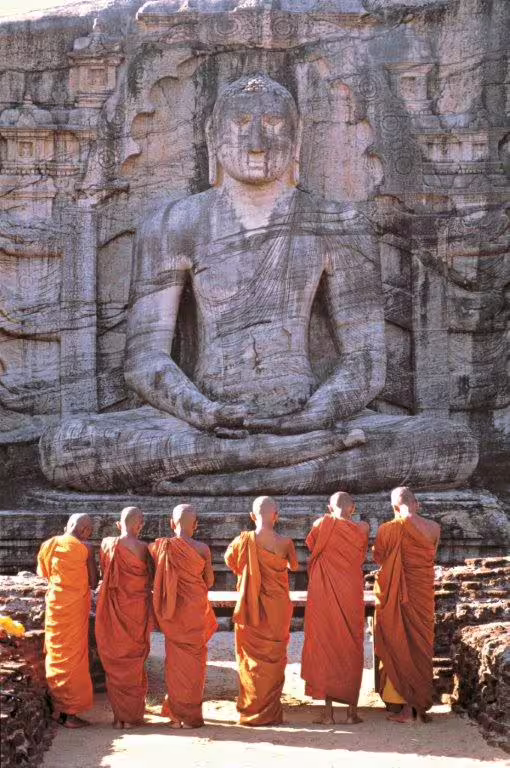 Buddhist monks in orange robes stand before an ancient rock carving of Buddha at Gal Vihara, Polonnaruwa, Sri Lanka.