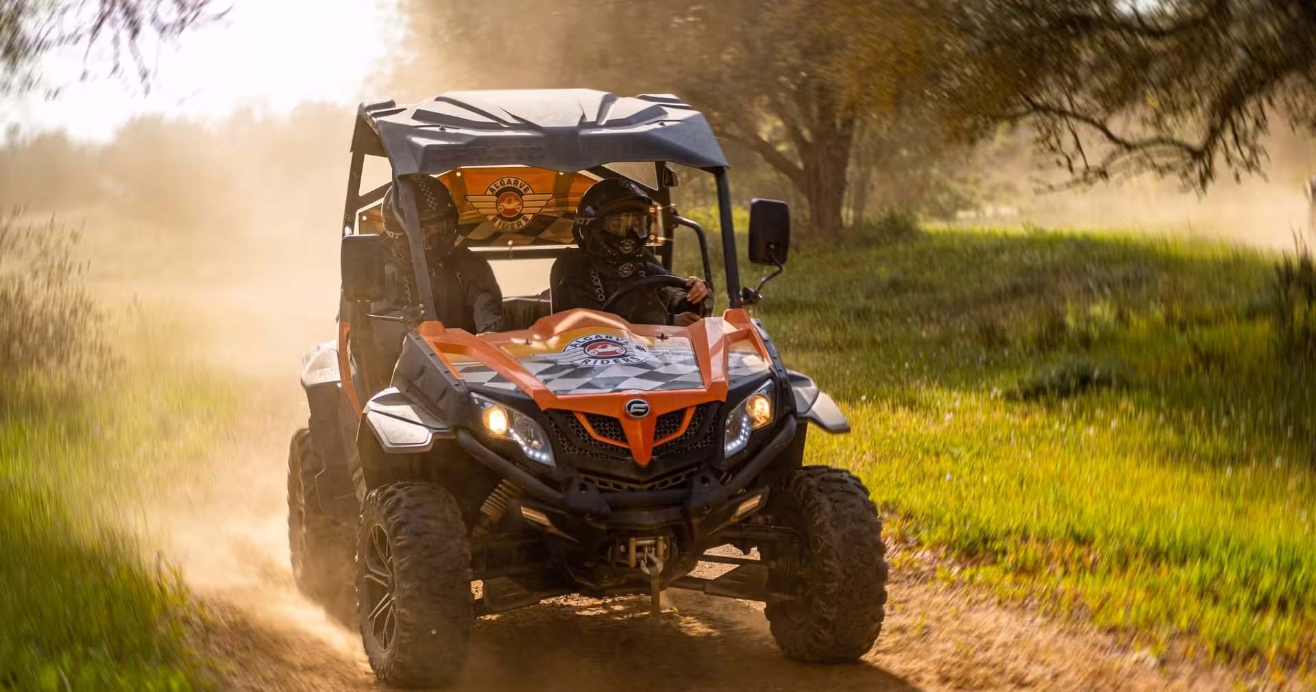 Two friends in helmets speeding through a sunny Algarve dirt track in an orange off-road buggy, kicking up dust and excitement