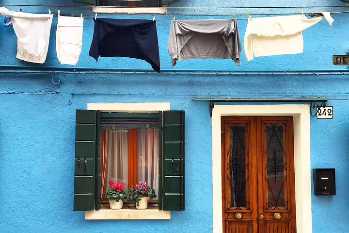 Vibrant blue house in Burano with laundry hanging and flowers in window, capturing the charm of a Grand Canal tour stop.