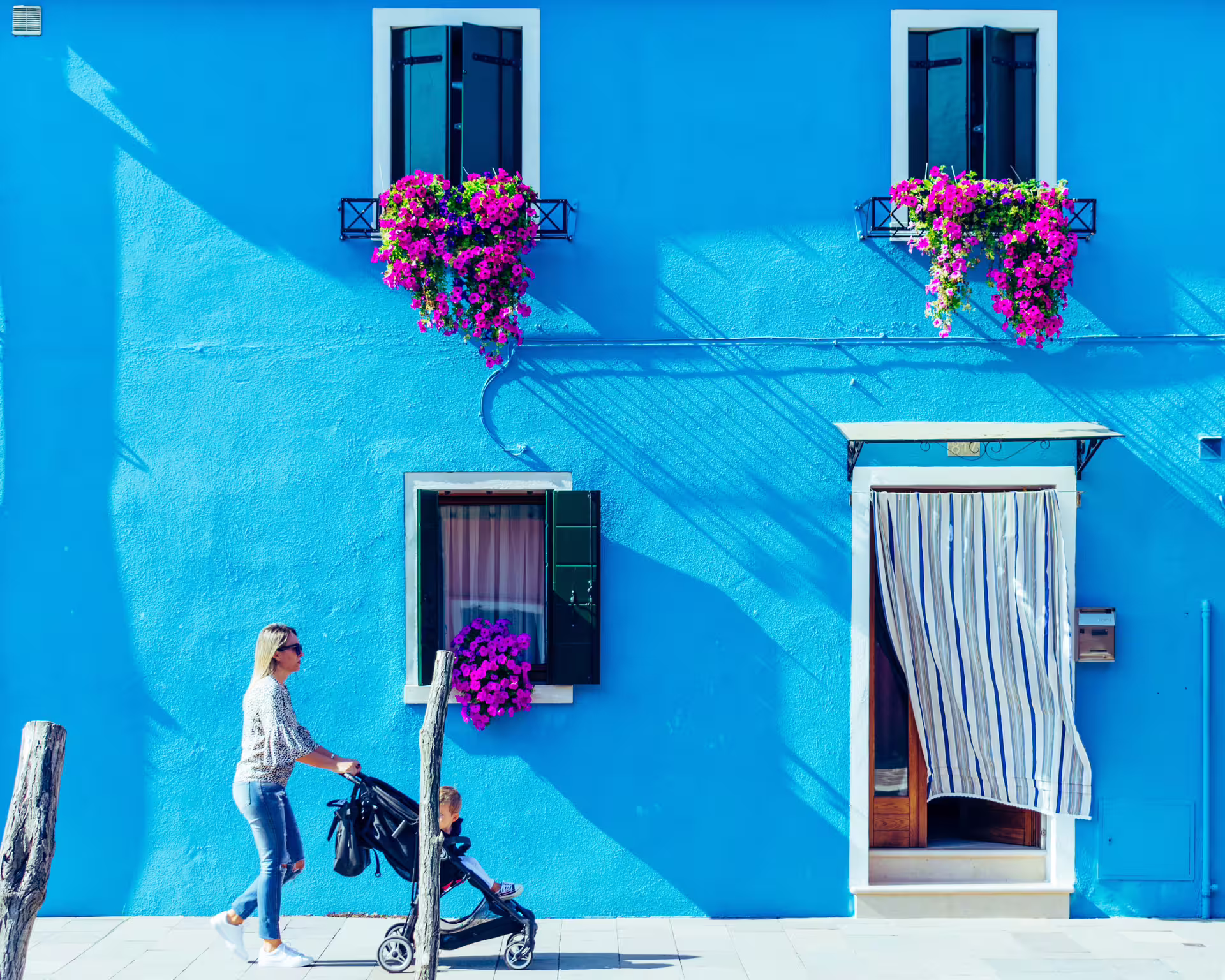 Woman with stroller passing a bright blue house adorned with pink flowers in Burano, ideal for a LimoBoat tour.