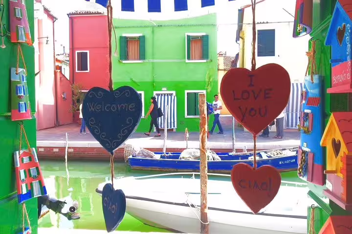 Heart-shaped signs by a canal in Burano, showcasing vibrant houses, part of the Murano and Burano water taxi tour.