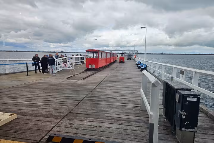 Scenic view of Busselton Jetty with a red train and tourists exploring the iconic wooden pier on a cloudy day.
