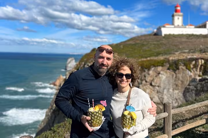 Couple enjoying tropical drinks at Cabo da Roca with the Atlantic Ocean and iconic lighthouse in the background on a Sintra tour.