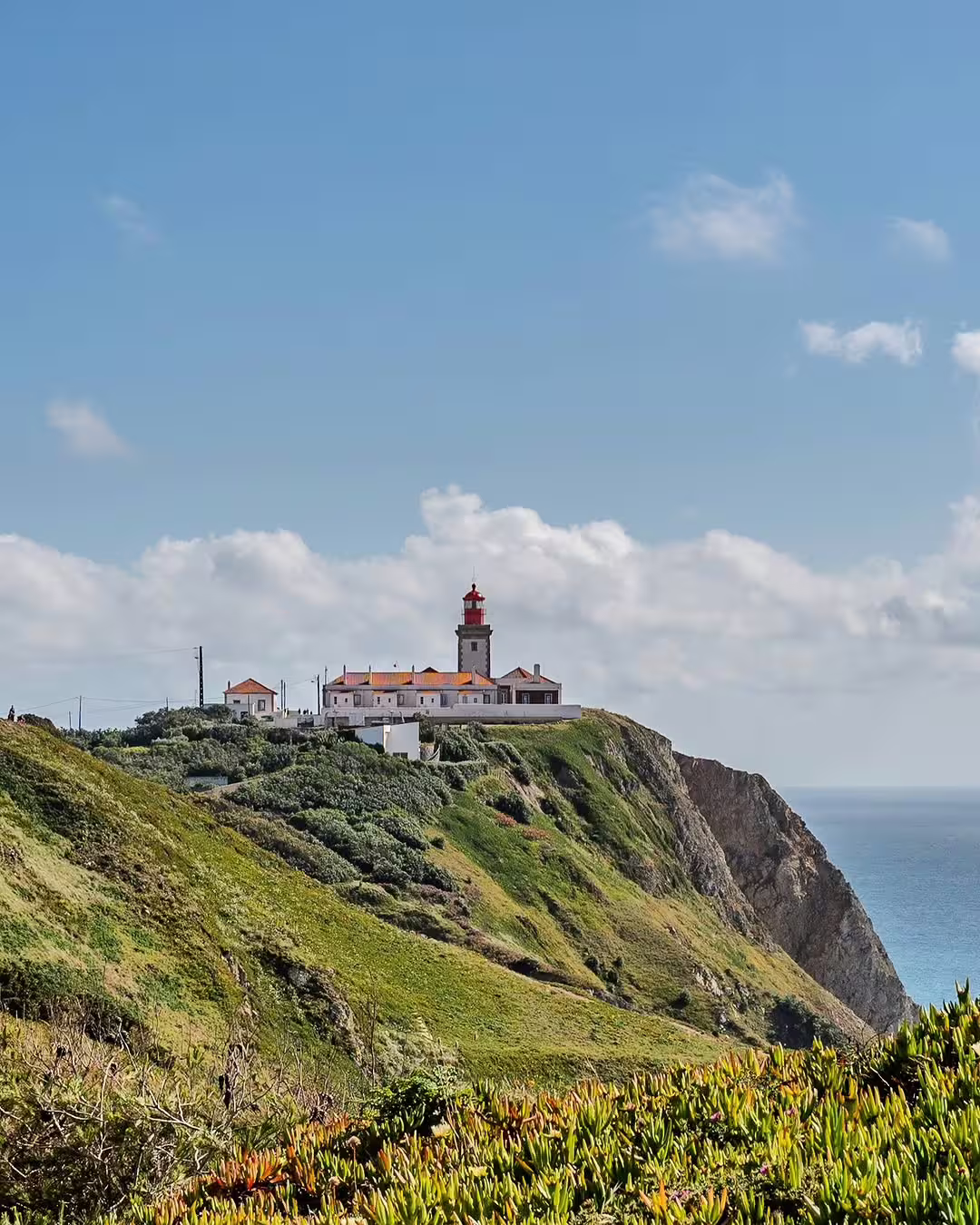 Scenic view of Cabo da Roca lighthouse perched on a cliff, surrounded by lush greenery and overlooking the Atlantic Ocean.