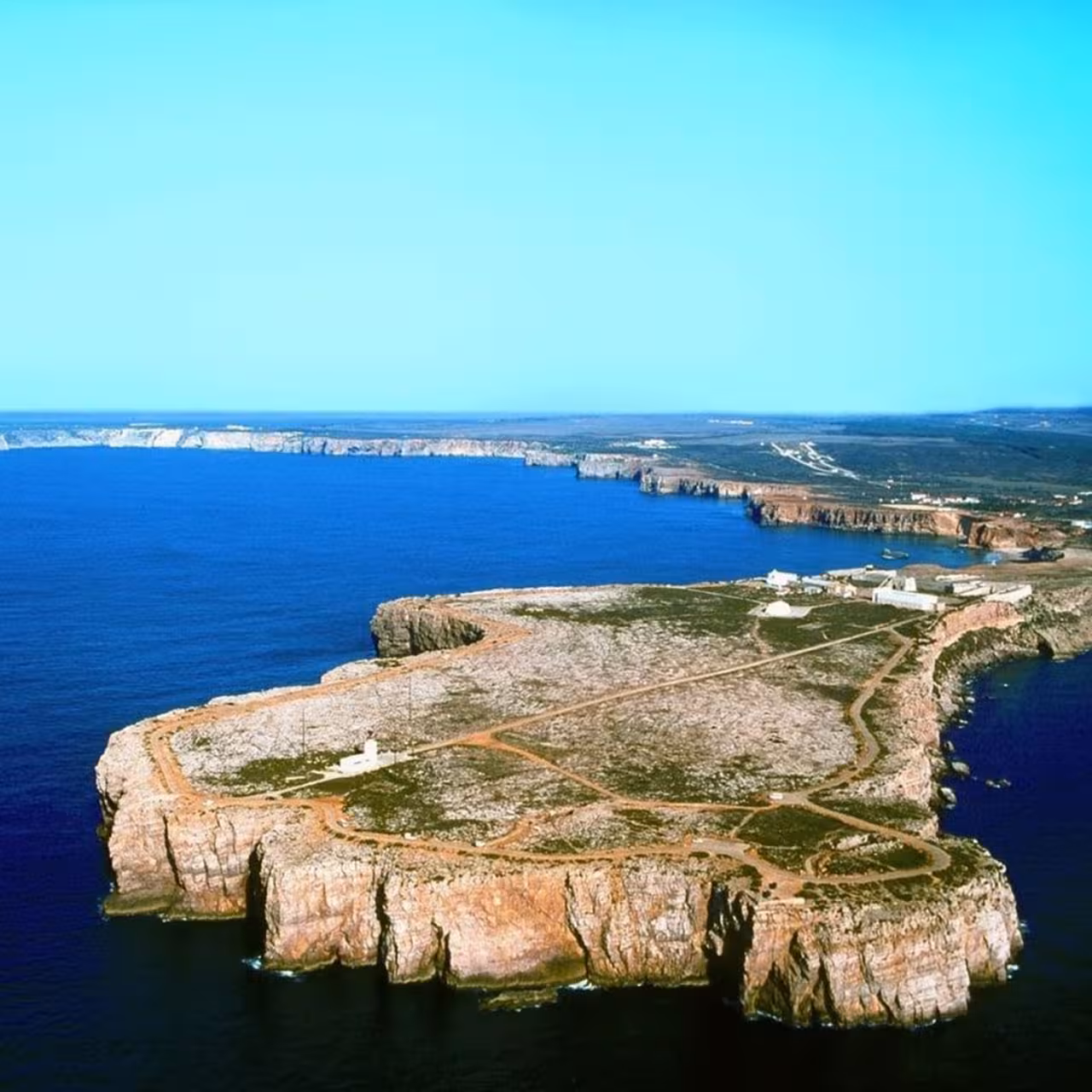 Aerial view of Cabo S. Vicente's rugged cliffs meeting the deep blue Atlantic Ocean under a clear sky.