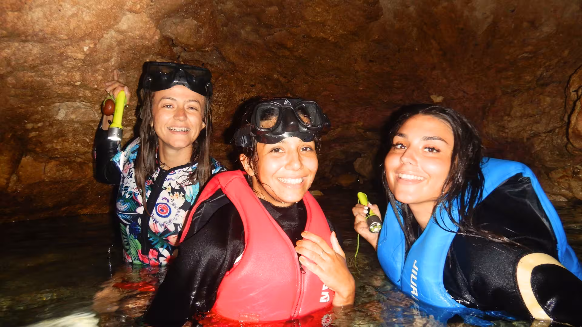 Three smiling friends snorkeling in a cave during Cagliari boat tour, equipped with life vests and flashlights.