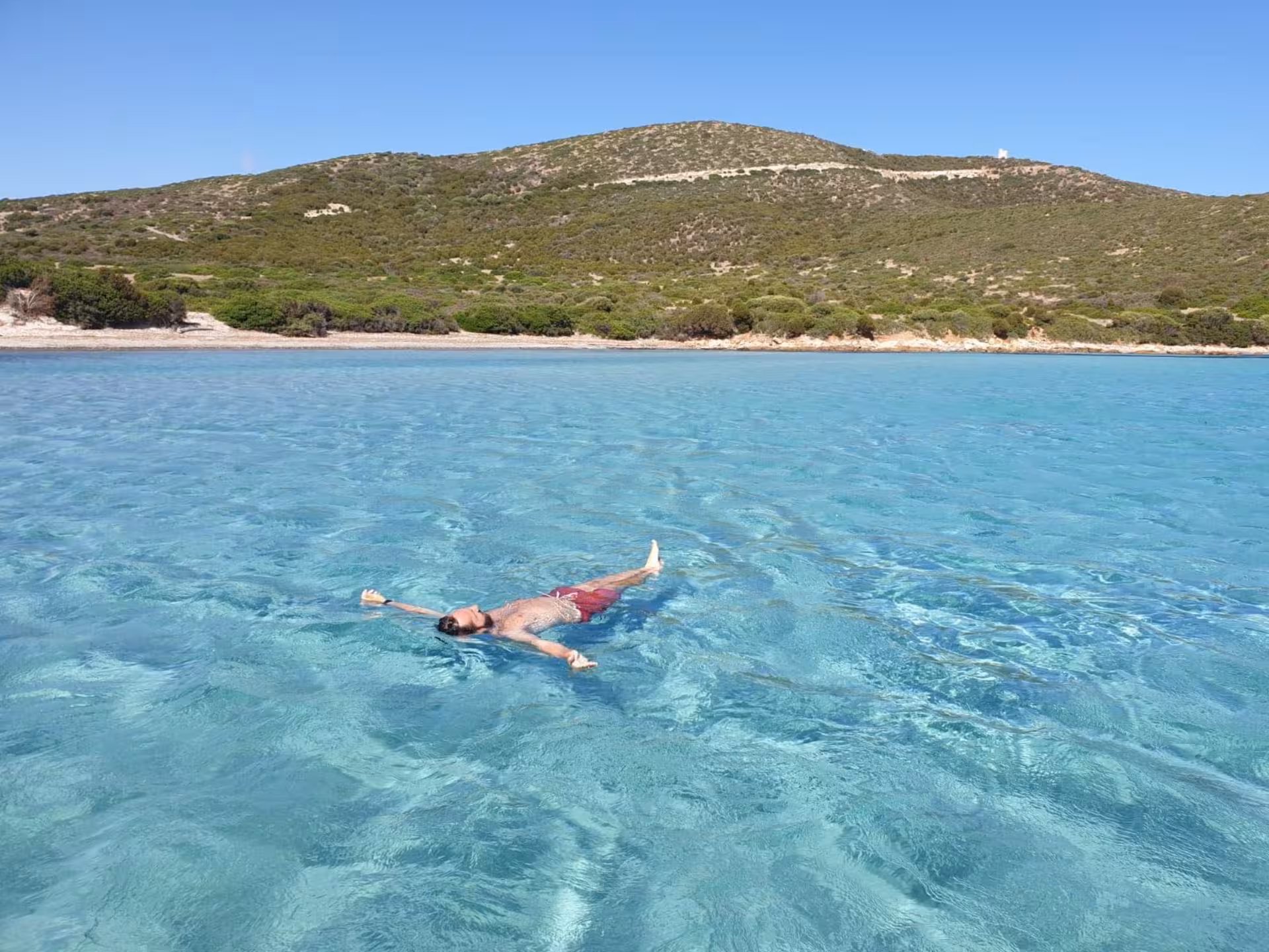 Person floating in serene, clear blue sea with picturesque hills in the background on Cagliari to Chia jeep tour.