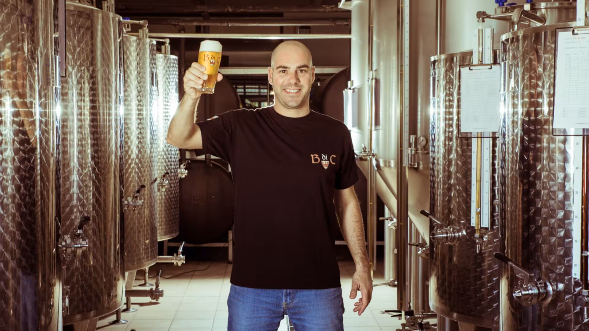 Smiling brewer holding a freshly poured beer in Cagliari's craft brewery tour, surrounded by shiny brewing tanks.