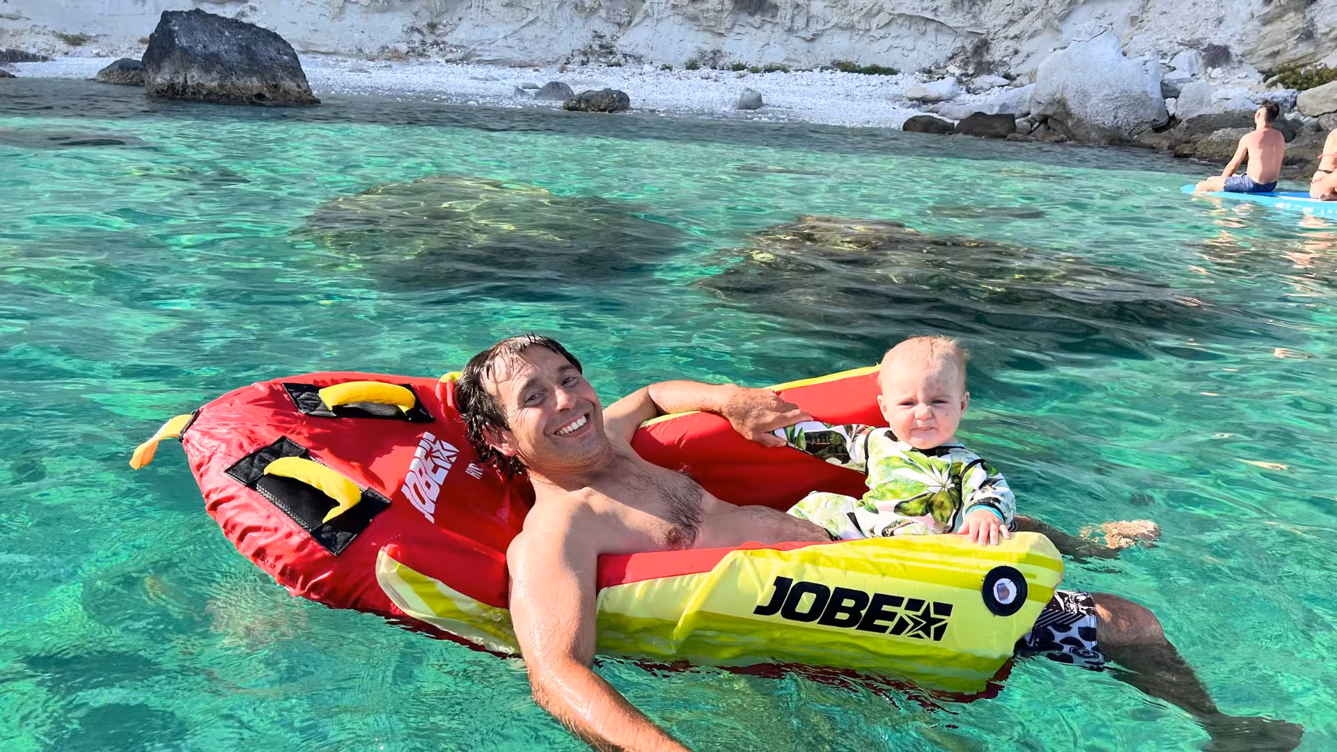 Smiling man and baby on inflatable raft in clear waters of Golfo degli Angeli, perfect for family water games.
