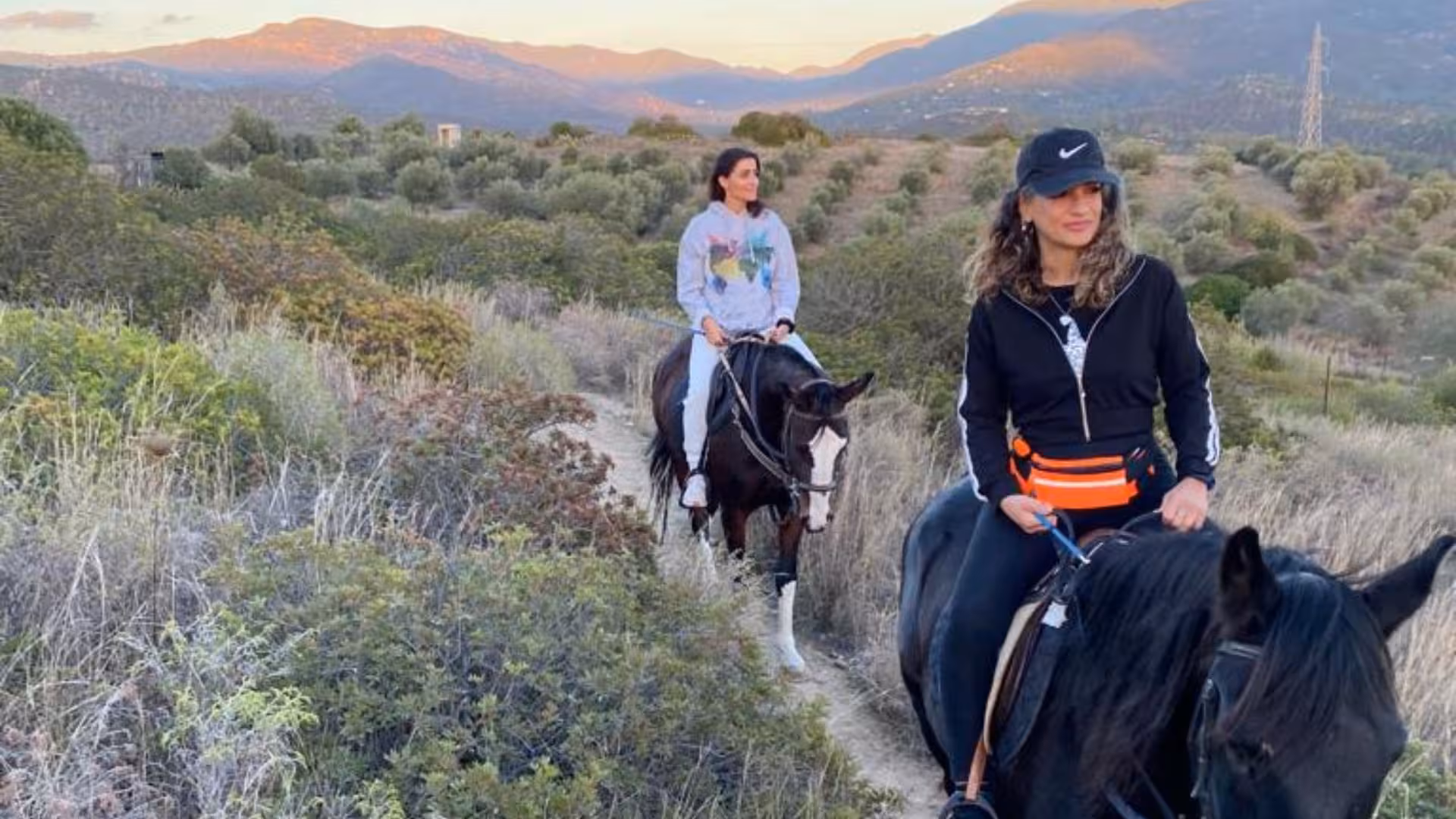Two riders explore Cagliari's scenic trails on horseback, surrounded by lush greenery and distant mountains.